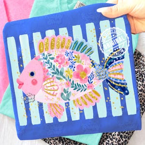 May include: A blue square decorative pillow featuring a whimsical pink fish design with floral patterns, gold accents, and a blue and white striped background. The pillow has the text "The Crafty Shop" and "Original Design".