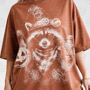以下が含まれることがあります： 錆色のTシャツで、カボチャ付きの魔女の帽子、ハート型のサングラス、チェック柄のバンダナを身に着けたアライグマの白いグラフィックプリントが施されています。横にはカボチャのバケツと「Trick or Treat」と書かれたカップが描かれています。