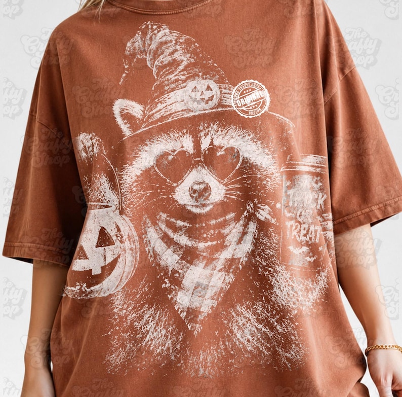 以下が含まれることがあります： 錆色のTシャツで、カボチャ付きの魔女の帽子、ハート型のサングラス、チェック柄のバンダナを身に着けたアライグマの白いグラフィックプリントが施されています。横にはカボチャのバケツと「Trick or Treat」と書かれたカップが描かれています。
