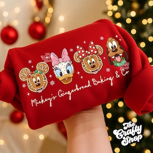 Pode incluir: Camisola vermelha com ilustrações de Mickey Mouse, Margarida, Minnie Mouse e Pluto em forma de biscoitos de gengibre. O texto "Mickey's Gingerbread Baking & Co" está impresso abaixo. O logotipo The Crafty Shop está no canto inferior direito.