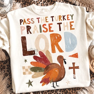 Peut inclure: Un sweat-shirt &agrave; col rond couleur cr&egrave;me avec le texte PASS THE TURKEY PRAISE THE LORD en lettres textur&eacute;es multicolores. Sous le texte se trouve un graphique repr&eacute;sentant une dinde brune aux plumes color&eacute;es, une petite croix brune et des &eacute;toiles.