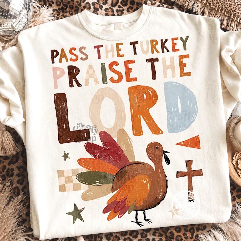 Peut inclure: Un sweat-shirt &agrave; col rond couleur cr&egrave;me avec le texte PASS THE TURKEY PRAISE THE LORD en lettres textur&eacute;es multicolores. Sous le texte se trouve un graphique repr&eacute;sentant une dinde brune aux plumes color&eacute;es, une petite croix brune et des &eacute;toiles.