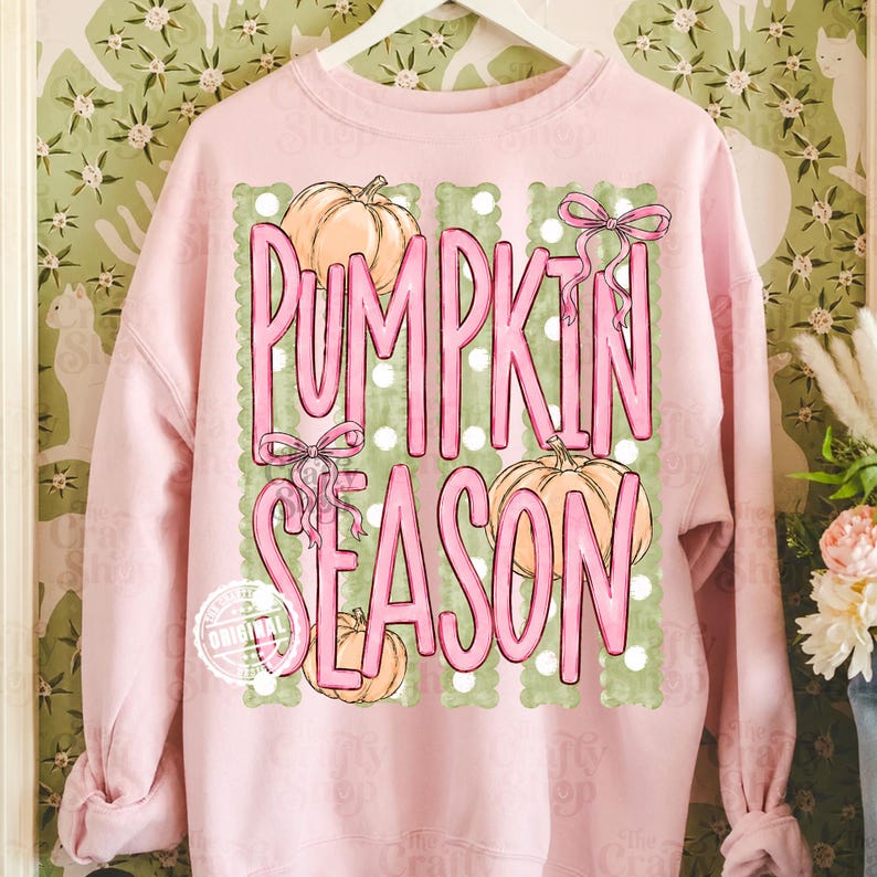 May include: A light pink long-sleeve sweatshirt featuring a graphic print with the text PUMPKIN SEASON in pink letters. The design includes three orange pumpkins, two pink bows, and vertical green stripes with white polka dots on a light pink background.