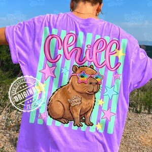 Op de afbeelding: Een paars T-shirt met een afbeelding van een capibara met een bril in de vorm van sterren en het woord "Chill" in roze letters. Het ontwerp bevat pastelgroene strepen en sterren. Het shirt heeft een relaxte pasvorm.