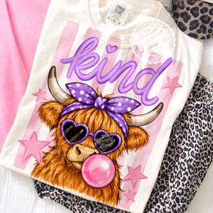 Pode incluir: Camiseta creme com um gráfico de uma vaca Highland usando óculos de sol em forma de coração e uma faixa de bolinhas, soprando uma bolha de chiclete. A palavra "kind" está escrita em roxo. Listras e estrelas rosa decoram a camiseta.