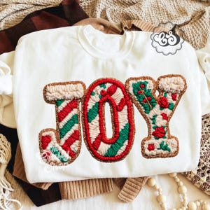 May include: White sweatshirt with the word "JOY" embroidered in red, green, and cream colors. The letters have a textured, festive design with gold trim, perfect for the holiday season.