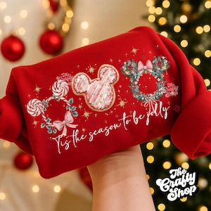 May include: A red sweatshirt featuring three Mickey Mouse head designs. One is a cookie, another a wreath, and the third a candy cane wreath. The text "Tis the season to be jolly" is printed below the designs. The Crafty Shop logo is in the bottom right.