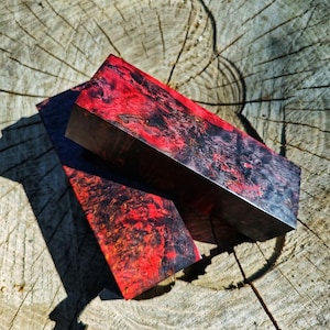 Pode incluir: Dois blocos retangulares de madeira estabilizada com um padrão marmoreado vibrante em vermelho profundo e preto. O veio da madeira apresenta uma textura complexa e rodopiante com alto contraste entre os tons carmesim e escuros.