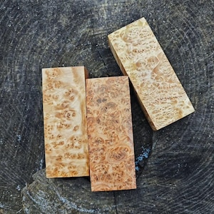 May include: Three rectangular wooden blocks with distinctive swirling grain patterns. The wood tones vary from light tan to reddish-brown, featuring intricate knots and textures. The blocks are arranged on a dark, textured surface.