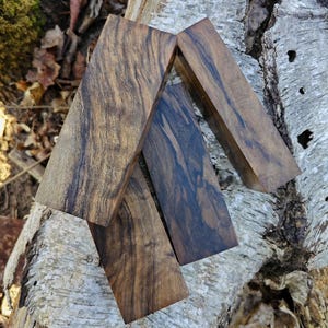 May include: Four rectangular blocks of dark brown wood with prominent black grain patterns. The pieces are arranged in a stacked, overlapping formation, showcasing the natural texture and varied dark streaks characteristic of the wood grain.