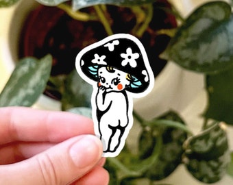 May include: A die-cut sticker featuring a cartoon character with a black mushroom cap adorned with white flowers. The character has a white body, light blue hair, and rosy red cheeks, depicted in a standing pose with one hand near its face.