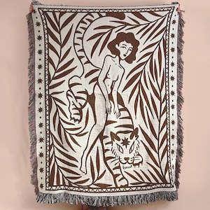 May include: A rectangular woven throw blanket featuring a brown and cream graphic design. The pattern depicts a figure sitting atop a tiger surrounded by large palm leaves and a border of stars. The blanket has a fringed edge on all sides.