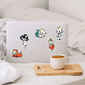 May include: Five die-cut stickers featuring cartoon babies in various costumes: a red devil, a black mushroom, a green mermaid, a strawberry, and an angel with blue wings. The stickers are arranged on the lid of a silver laptop computer.
