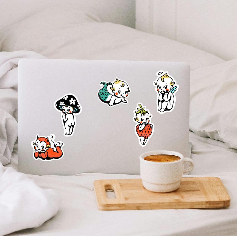 May include: Five die-cut stickers featuring cartoon babies in various costumes: a red devil, a black mushroom, a green mermaid, a strawberry, and an angel with blue wings. The stickers are arranged on the lid of a silver laptop computer.