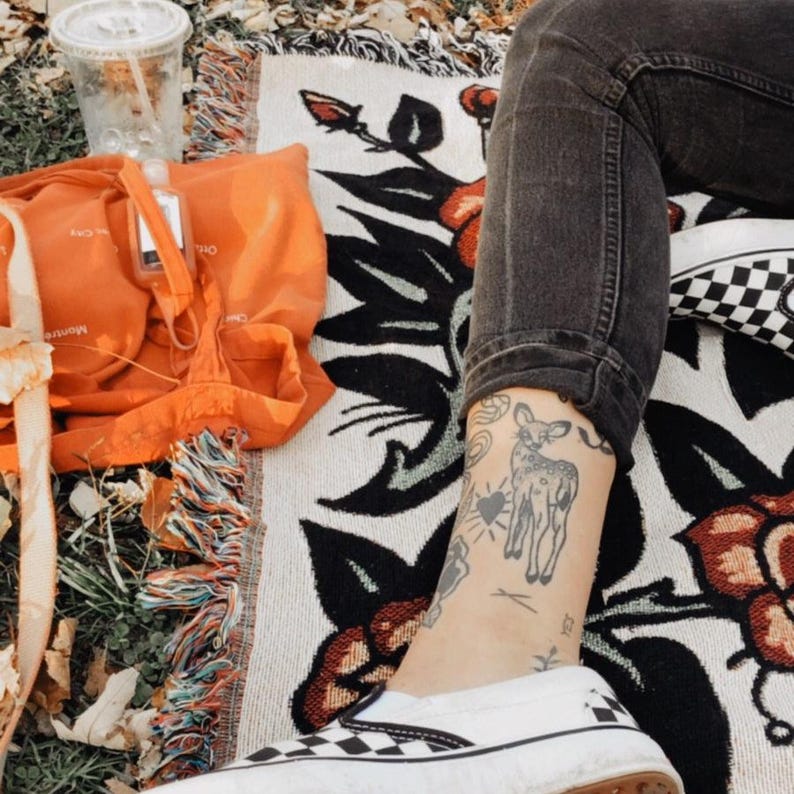 May include: A woven blanket featuring a black and red floral pattern, an orange tote bag, a clear plastic cup, and a person wearing black jeans and black and white checkered slip-on sneakers with a deer tattoo on their lower leg.
