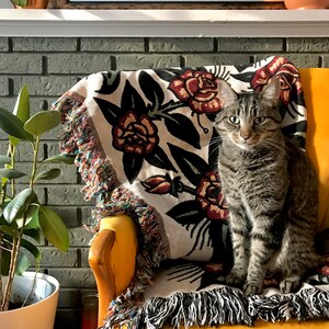 May include: A woven throw blanket featuring a floral pattern of red roses and dark green leaves on a cream background, finished with multicolored fringe edges. A brown tabby cat sits on the blanket, which is draped over a yellow chair.