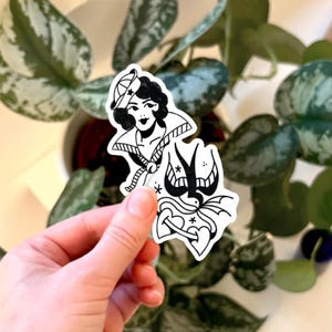 May include: Black and white sticker with a vintage tattoo design. The sticker shows a woman in a sailor hat and a swallow with a heart and stars. The design is in the style of traditional tattoo art.