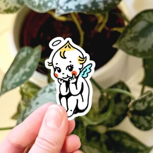 May include: A die-cut sticker featuring a cartoon cherub with yellow hair, a small halo, and light blue wings. The character has rosy red cheeks, large eyes, and is depicted in a seated position with hands resting near the chin, outlined in black.