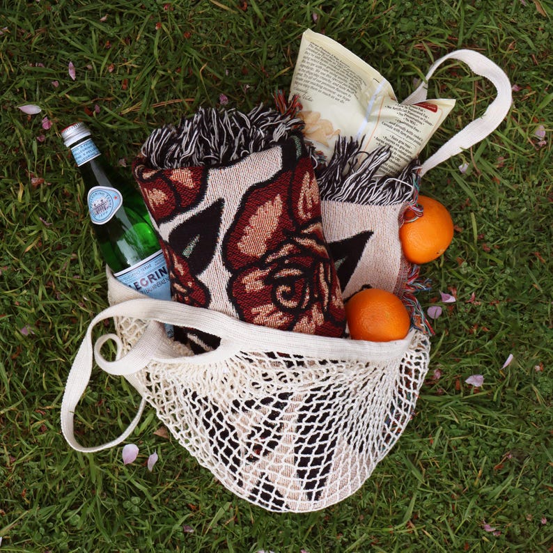 May include: A cream-colored cotton string market bag containing a woven blanket with a dark red and black rose pattern, a glass bottle of San Pellegrino sparkling water, a bag of snacks, and two fresh oranges nestled inside the mesh.