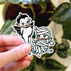 May include: Two Halloween-themed stickers featuring a cute cartoon vampire and a mummy. The vampire is wearing a black cape and has yellow hair. The mummy is wrapped in white bandages and has a blue face.