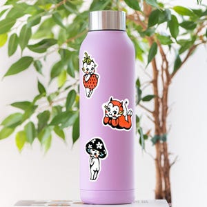 May include: A light purple stainless steel water bottle with a silver cap, decorated with three stickers featuring vintage-style cartoon characters: a child dressed as a strawberry, a red devil character, and a child standing under a black mushroom.