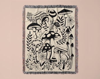 May include: A woven throw blanket featuring a cream-colored base with a repeating black graphic print of various mushrooms, fungi, and small flowers. The rectangular textile has a decorative fringed edge along the top and bottom borders.