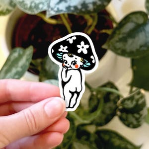 May include: A die-cut sticker featuring a cartoon character with a black mushroom cap adorned with white flowers. The character has a pale white body, rosy red cheeks, and light blue hair, depicted in a standing pose with a hand near the face.