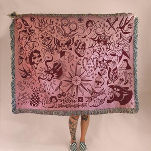 May include: A woven throw blanket in shades of pink and dark red, featuring a variety of tattoo-style illustrations including a panther, skull, heart, sun, and cherubs. The text "how lucky we are" is printed in the bottom left corner with fringe.