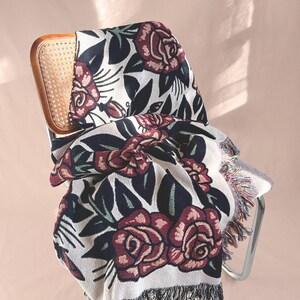 May include: A woven throw blanket featuring a repeating pattern of dark red roses with black leaves and stems on a cream-colored background. The blanket has a textured knit construction and is finished with decorative fringe along the bottom edge.
