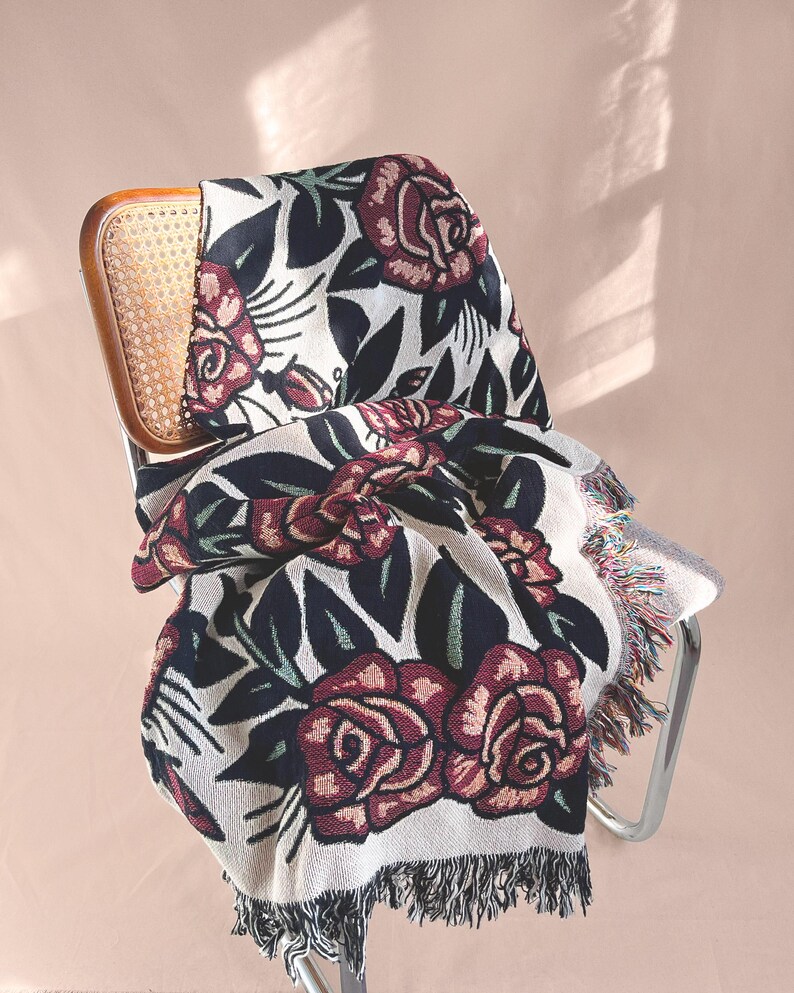May include: A woven throw blanket featuring a repeating pattern of dark red roses with black leaves and stems on a cream-colored background. The blanket has a textured knit construction and is finished with decorative fringe along the bottom edge.