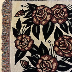 May include: A woven throw blanket featuring a repeating pattern of dark red roses with black leaves on a cream-colored background. The edges of the blanket are finished with a multicolored fringe made of various yarn colors.