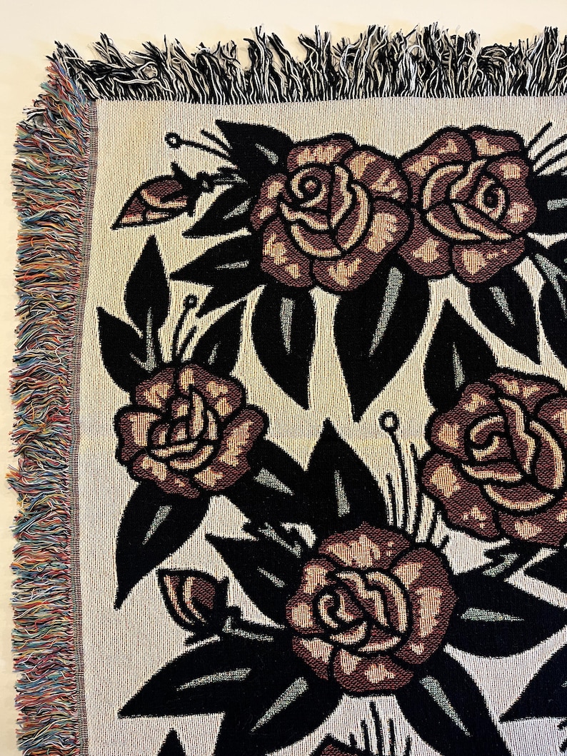 May include: A woven throw blanket featuring a repeating pattern of dark red roses with black leaves on a cream-colored background. The edges of the blanket are finished with a multicolored fringe made of various yarn colors.