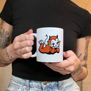 May include: White ceramic mug featuring a cartoon illustration of a red devil with yellow horns and a white halo. The devil is lying on its side with its head resting on its hand.