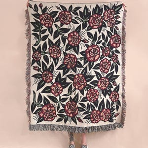 May include: A rectangular woven throw blanket featuring a repeating pattern of dark red roses with deep green leaves on a cream-colored background. The blanket has a textured surface and is finished with decorative fringe along the vertical edges.
