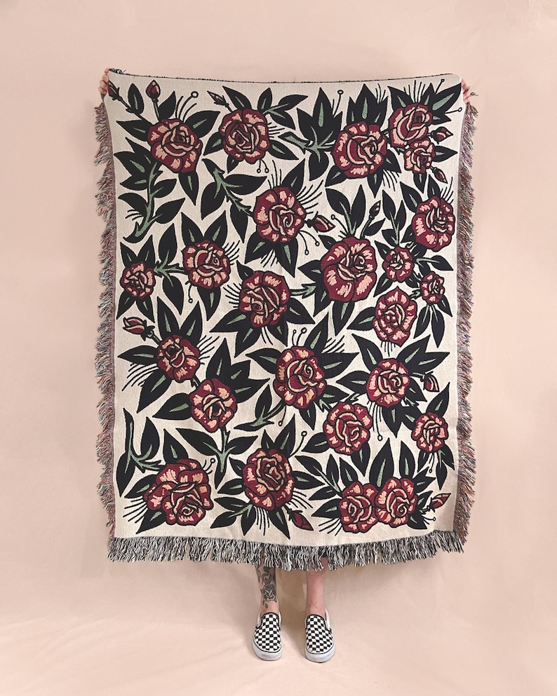May include: A rectangular woven throw blanket featuring a repeating pattern of dark red roses with deep green leaves on a cream-colored background. The blanket has a textured surface and is finished with decorative fringe along the vertical edges.