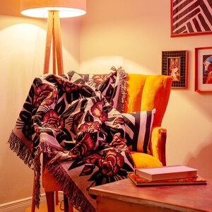 May include: A woven throw blanket featuring a repeating pattern of red roses with black leaves and outlines on a cream background, finished with fringed edges, draped over a bright orange upholstered armchair in a room with warm lighting.
