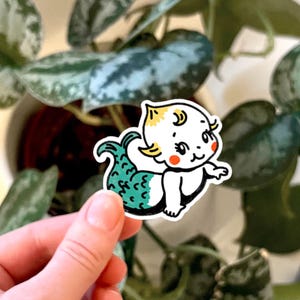 May include: A die-cut sticker featuring a cartoon baby mermaid with pale skin, yellow hair, and bright red cheeks. The mermaid has a teal tail with dark green scale details and black outlines, presented in a clean, graphic illustration style.