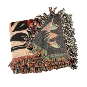 May include: A woven throw blanket featuring a botanical pattern with large black leaves on a tan background. The reverse side displays a dark green and multicolored floral design. The edges are finished with long, multicolored fringe.