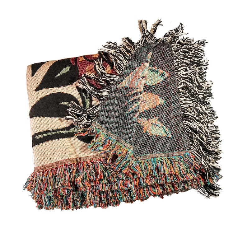 May include: A woven throw blanket featuring a botanical pattern with large black leaves on a tan background. The reverse side displays a dark green and multicolored floral design. The edges are finished with long, multicolored fringe.