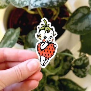 May include: A die-cut sticker featuring a vintage-style illustration of a baby with blonde hair and rosy cheeks, wearing a red strawberry costume with black seeds. The character has a green stem on its head and is holding the strawberry body.