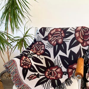 May include: A woven throw blanket featuring a repeating pattern of dark red roses with black leaves on a light cream background. The blanket has a fringed edge with multicolored threads and is draped over a chair.