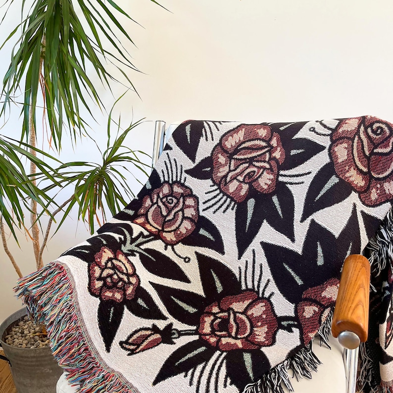 May include: A woven throw blanket featuring a repeating pattern of dark red roses with black leaves on a light cream background. The blanket has a fringed edge with multicolored threads and is draped over a chair.