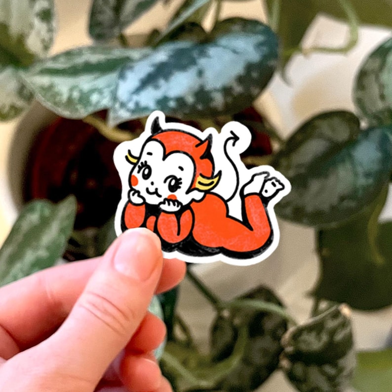 May include: A die-cut sticker featuring a cartoon character with white skin, red cheeks, and yellow hair accents, dressed in a bright orange devil costume with horns and a tail, resting on its stomach with hands propped under its chin.