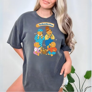 Puede incluir: Una camiseta gris oscuro con un gráfico de los osos Berenstain. Los osos llevan ropa y sostienen una olla de miel. El texto "The Berenstain Bears" está por encima de los osos.