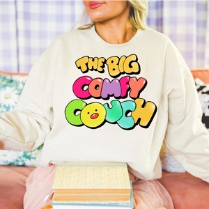 K&ouml;nnte beinhalten: Ein wei&szlig;er Pullover mit dem Text "THE BIG COMFY COUCH" in bunten Blasenbuchstaben. Die Buchstaben sind schwarz umrandet und haben einen 3D-Effekt. Ein gelbes Comic-Gesicht mit roter Nase befindet sich in der Mitte des Wortes "COUCH".