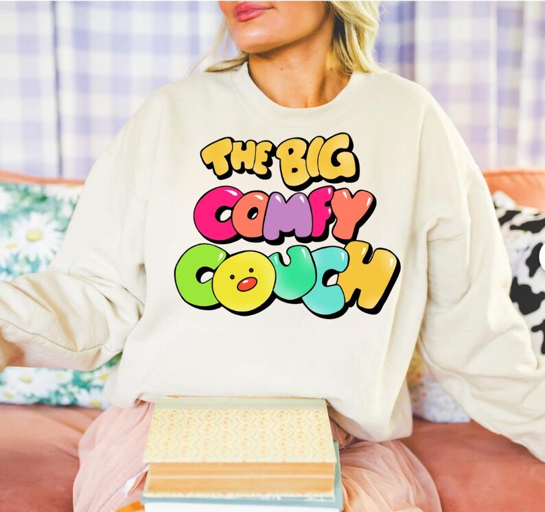 K&ouml;nnte beinhalten: Ein wei&szlig;er Pullover mit dem Text "THE BIG COMFY COUCH" in bunten Blasenbuchstaben. Die Buchstaben sind schwarz umrandet und haben einen 3D-Effekt. Ein gelbes Comic-Gesicht mit roter Nase befindet sich in der Mitte des Wortes "COUCH".