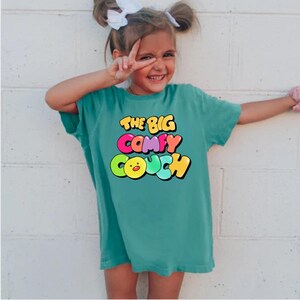 K&ouml;nnte beinhalten: Ein t&uuml;rkisfarbenes T-Shirt mit einer bunten Grafik, auf der "The Big Comfy Couch" steht. Die Grafik zeigt ein gelbes K&uuml;ken mit einem rosa Schnabel und einer regenbogenfarbenen Schrift.
