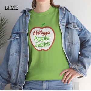 Könnte beinhalten: Hellgrünes T-Shirt mit dem Kellogg's Apple Jacks-Logo in roter und weißer Apfelform. Das Shirt wird mit einer blauen Jeansjacke und Jeans getragen. Das Wort "LIME" befindet sich in der oberen linken Ecke.