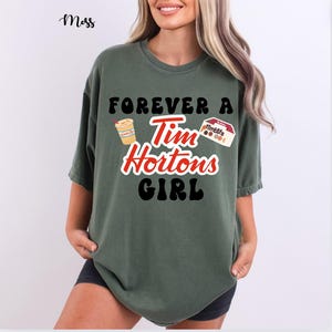 Puede incluir: Camiseta verde musgo con el texto "FOREVER A Tim Hortons GIRL" en negro y rojo. El diseño incluye ilustraciones de una taza de café y una caja de Timbits. La camiseta tiene mangas cortas.