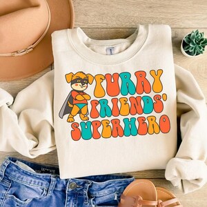 May include: A cream-coloured sweatshirt with a retro-style graphic of a cartoon dog wearing a superhero costume. The text "Furry Friends' Superhero" is in colourful, bubble-like letters.